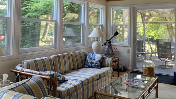A sunroom with a wooden ceiling, large windows, a striped sofa, a glass coffee table, and a telescope.