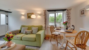 Una sala de estar acogedora con un sofá verde, una mesa de comedor y sillas. Hay una ventana con cortinas, un jarrón con flores y un cuadro en la pared.