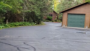 Ein gepflasterter Weg führt zu einer Hütte mit einer grünen Garagenöffnung, umgeben von dichtem Wald.