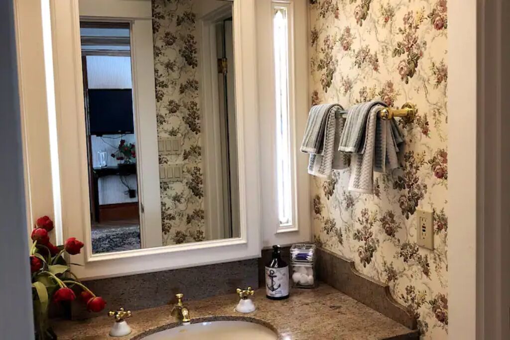 Ein Badezimmer mit blumigem Tapetenmuster, einem Waschbecken mit goldfarbenen Armaturen und einem Spiegel, der einen Raum mit einem Fenster und einer Vase mit roten Blumen reflektiert.