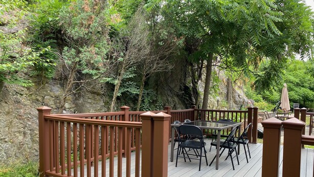Une terrasse en bois avec une table et des chaises en verre, entourée d’une balustrade en bois et d’une végétation luxuriante.