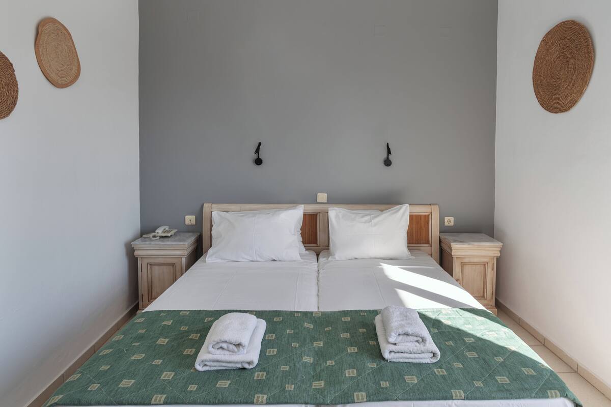 Una cama doble con sábanas blancas, dos mesitas de noche, una colcha estampada verde y dos toallas dobladas.