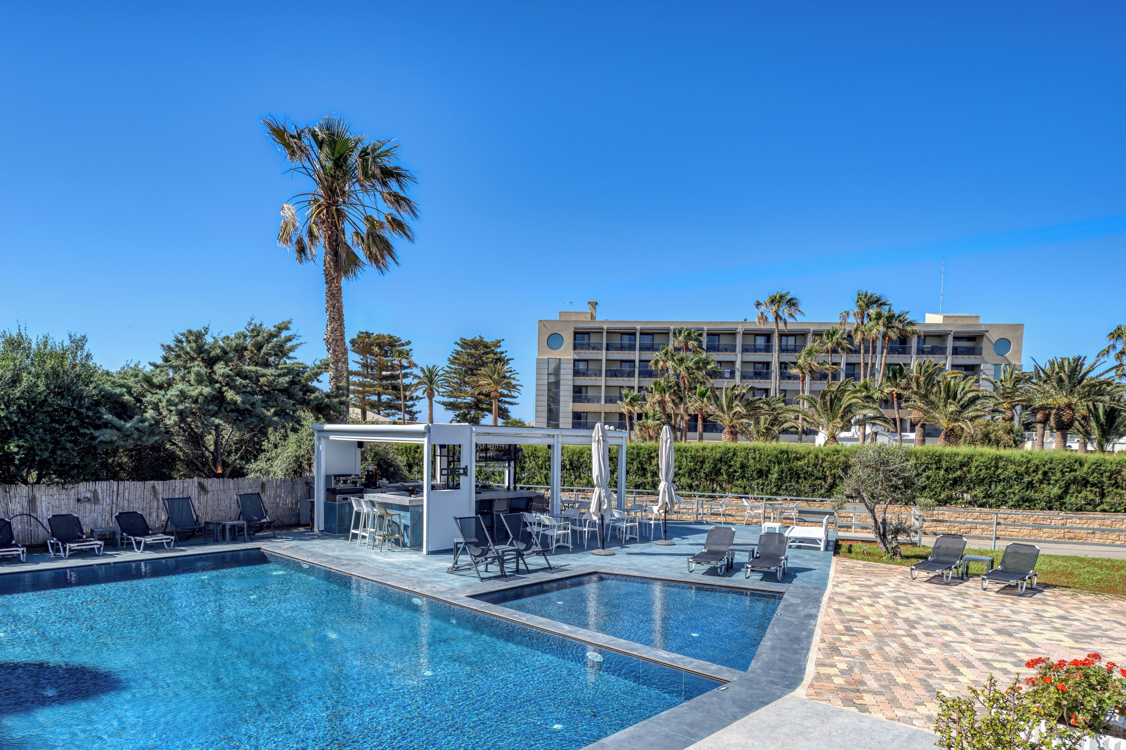 A swimming pool with lounge chairs and umbrellas, a bar area, and a building with multiple floors and balconies in the background.