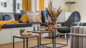 Una sala de estar moderna con un sofá, una mesa de centro y una silla. Hay una lámpara, un florero con plantas secas y una botella sobre la mesa de centro.