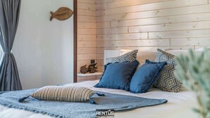 Un dormitorio con pared de tablones de madera, una cama con almohadas azules y estampadas, una almohada a rayas y una pequeña estantería con objetos decorativos.