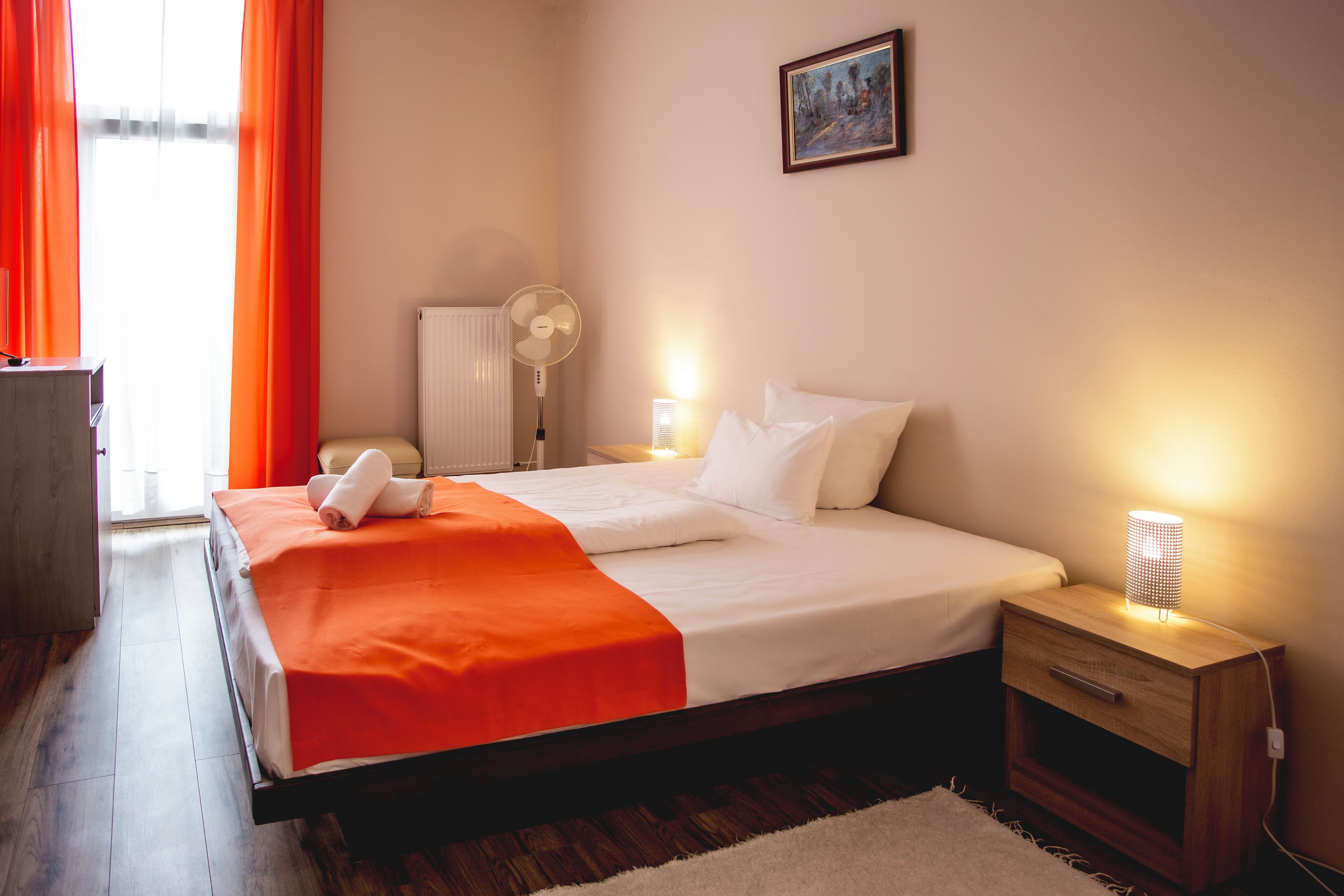 Una cama bien tendida con ropa de cama blanca y naranja, una mesita de noche con una lámpara y un radiador en una habitación con suelo de madera.