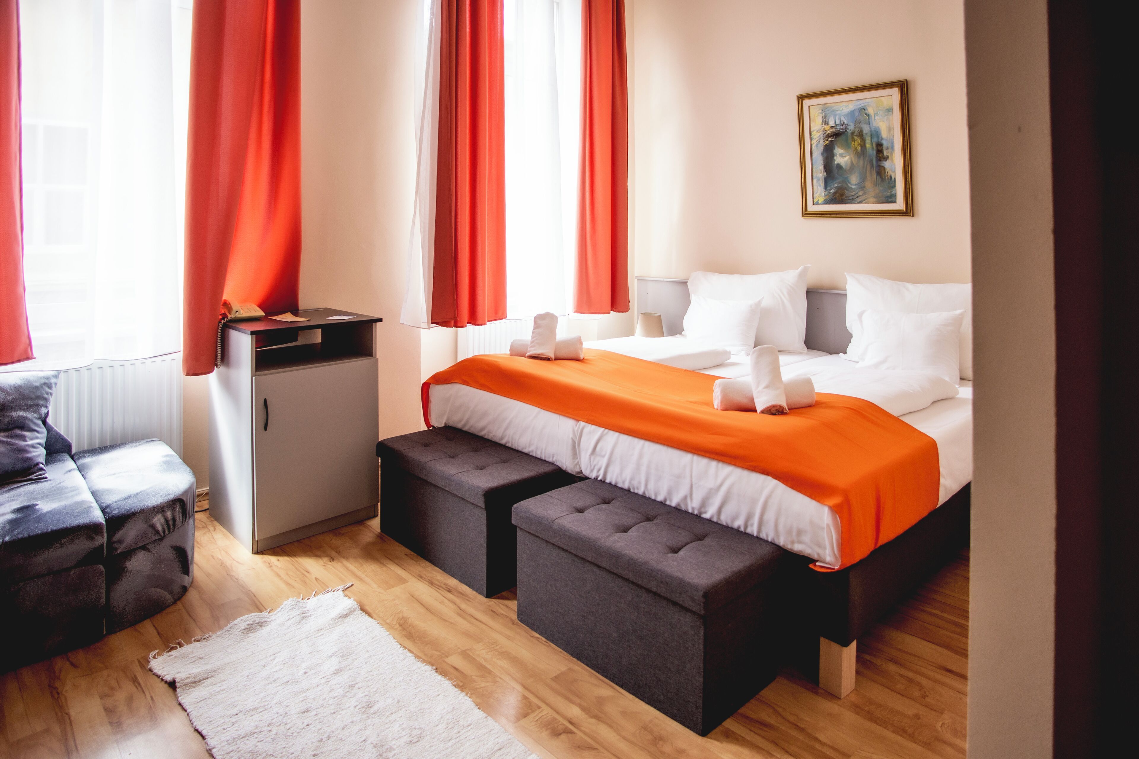 A hotel room with a bed, two ottomans, a desk, a chair, and a painting on the wall.