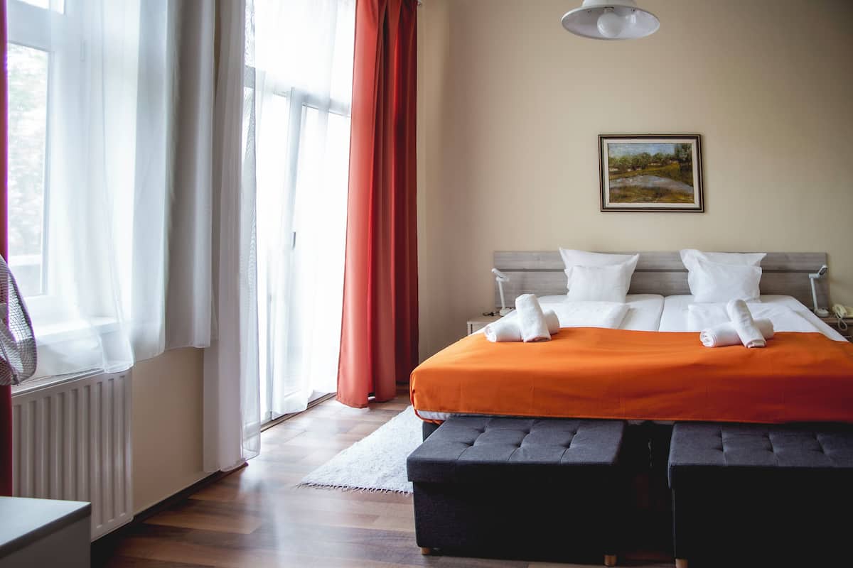 Ein ordentlich bezogenes Bett mit weißen Leinen und einer orangefarbenen Decke, eine graue gepolsterte Bank und ein Gemälde an der Wand.