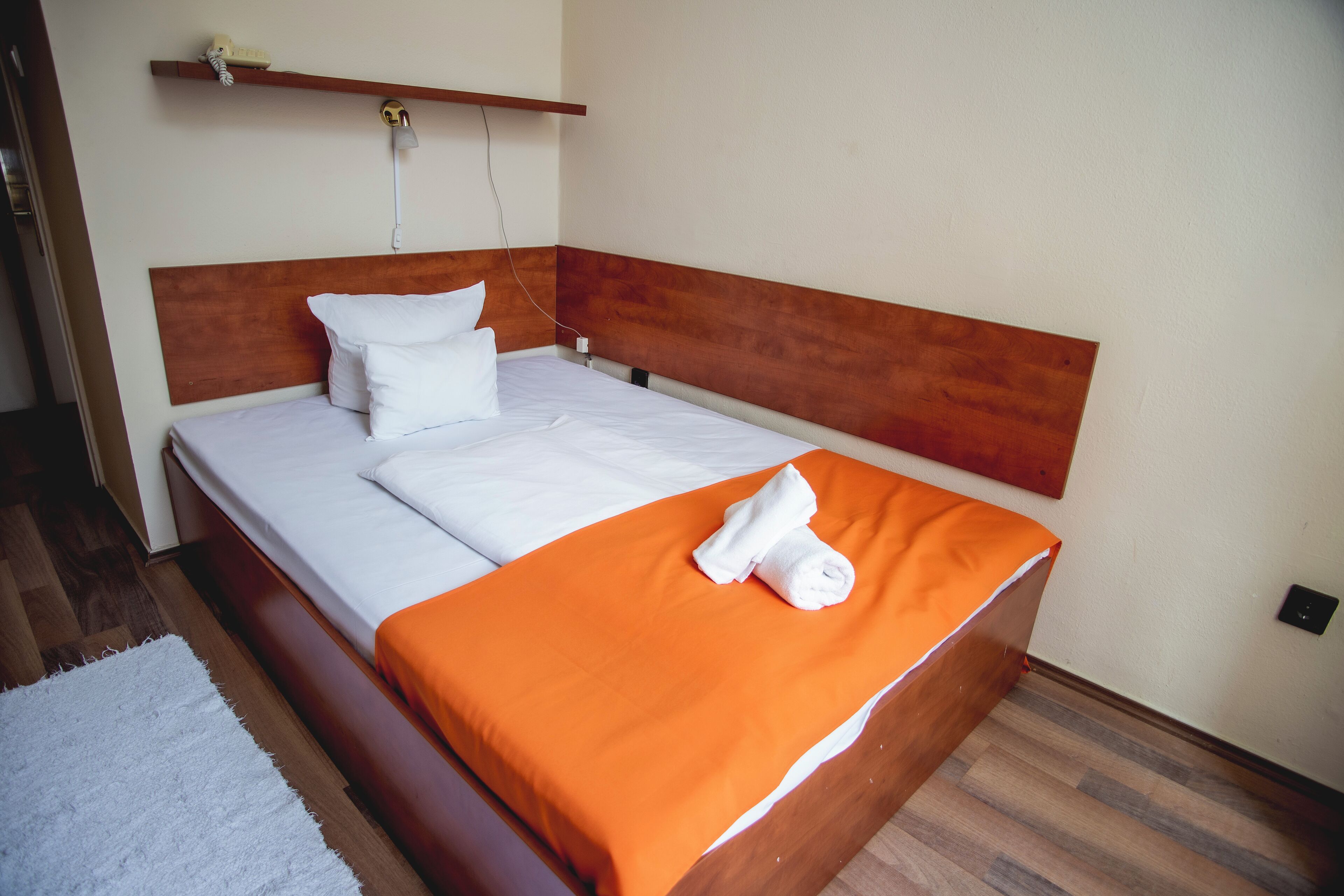 A neatly made bed with white linens and an orange blanket, a wooden headboard, and a white wall with a power outlet.
