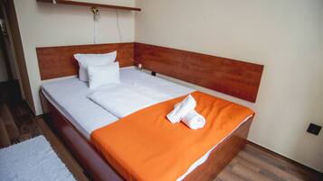 A neatly made bed with white linens and an orange blanket, a wooden headboard, and a white wall with a power outlet.