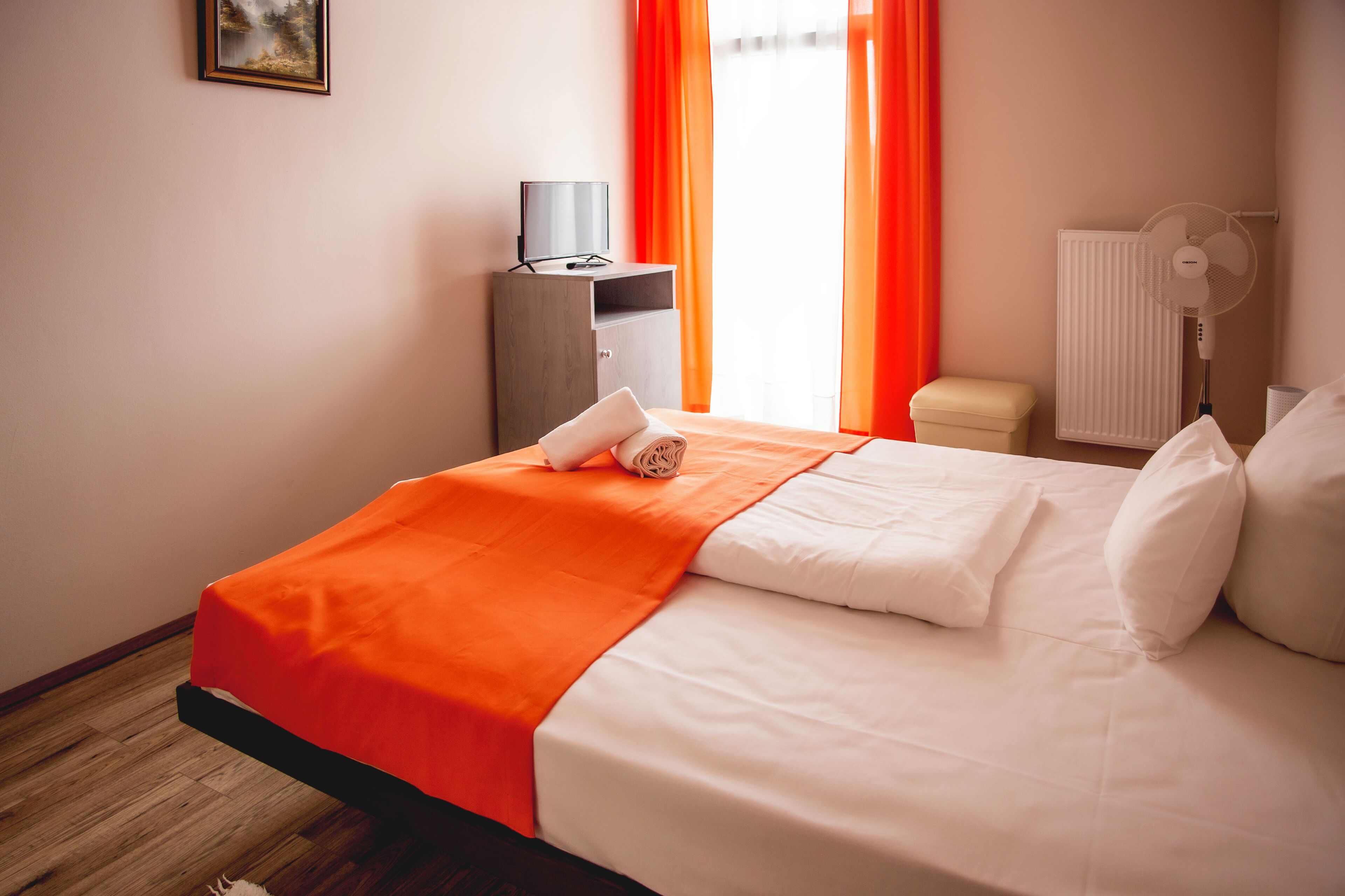 Una cama bien hecha con ropa de cama blanca y naranja, una mesita de noche con un televisor y un radiador.