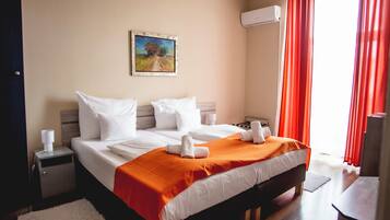A hotel room with a bed, a nightstand, a painting on the wall, and an air conditioning unit.