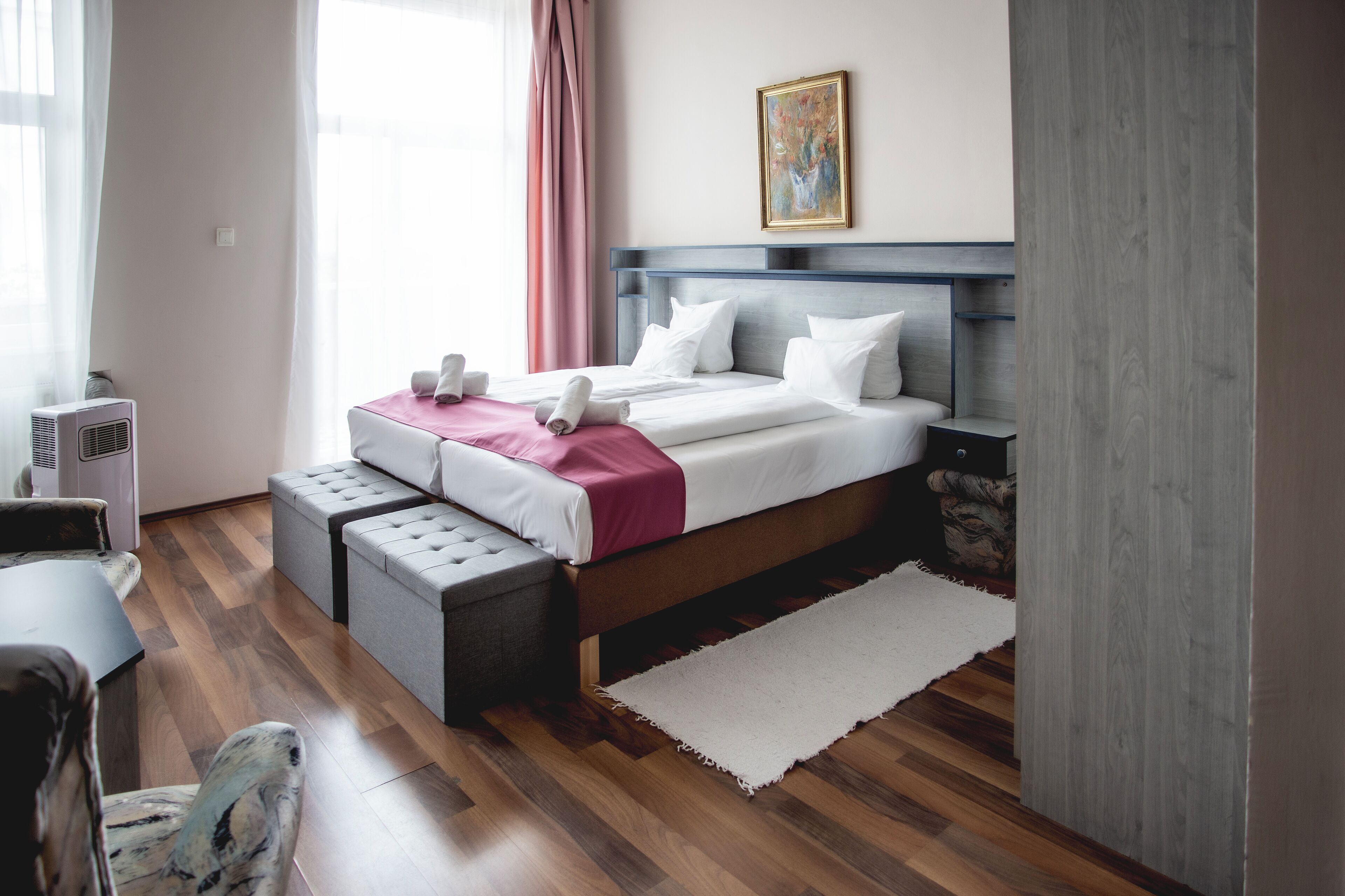 A modern hotel room with a wooden floor, a bed with white linens, a grey headboard, and a painting on the wall.