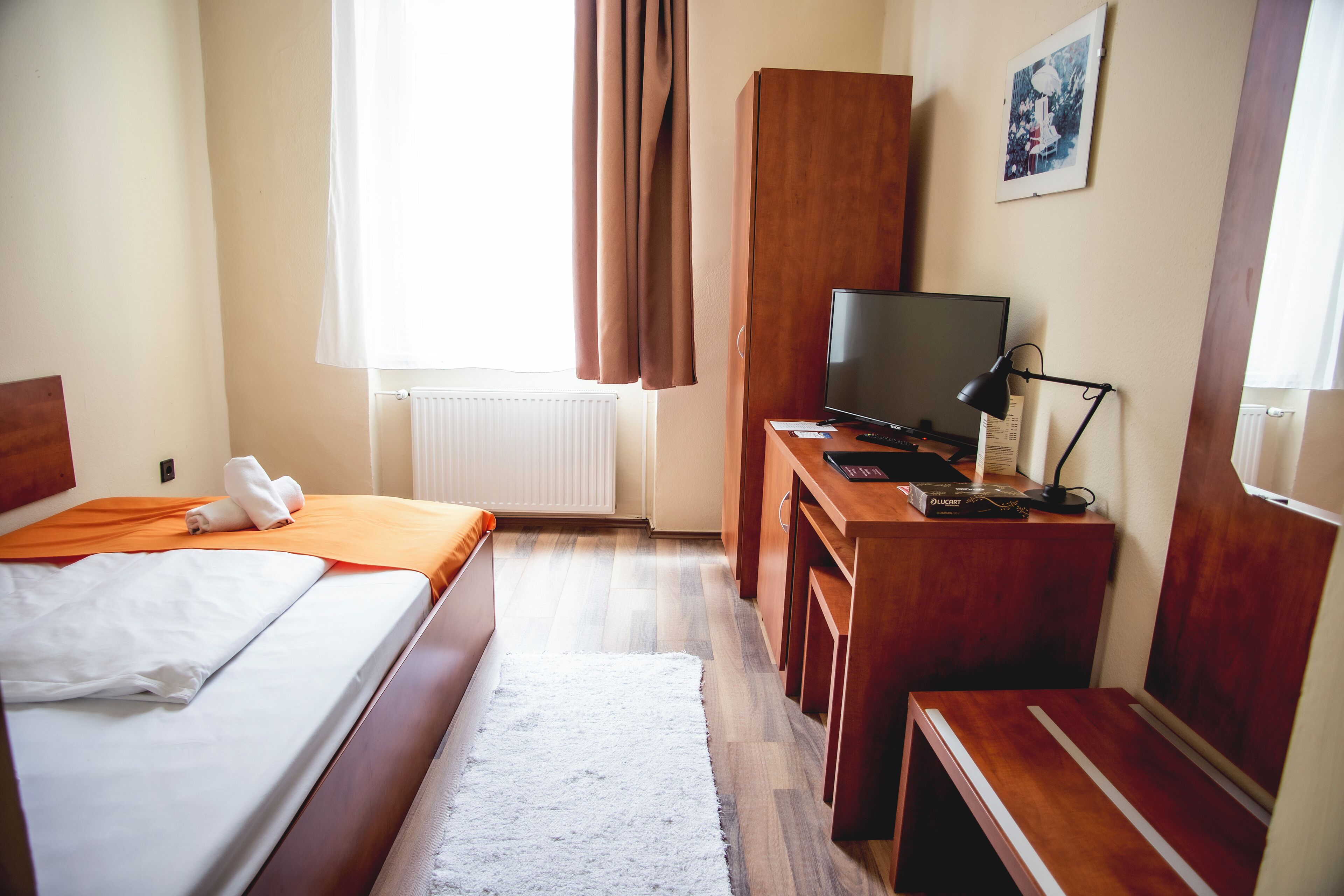 Quarto de hotel com cama, escrivaninha com computador, televisão e radiador.