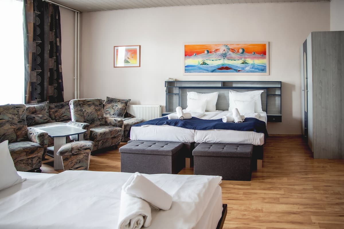 Ein Hotelzimmer mit Bett, zwei Sesseln, einem kleinen Tisch und einer Bank. An der Wand hängen zwei Gemälde.