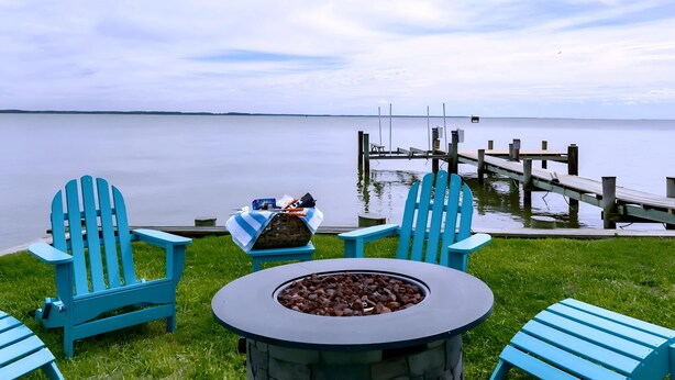 A lakeside seating area with a fire pit, two Adirondack chairs, and a dock extending into the water. - Waterfront Tilghman Retreat with Dock, Fire Pit & Sunsets (Tilghman Island)