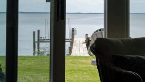 A view from a room with a sliding glass door to a deck overlooking a body of water, with a chair and a table visible. - Waterfront Tilghman Retreat with Dock, Fire Pit & Sunsets (Tilghman Island)