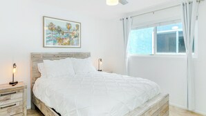 A bedroom with a wooden headboard, a white bedspread, two bedside tables with lamps, a ceiling fan, a window with curtains, and a painting on the wall.