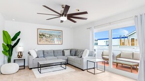 A modern living room with a grey sectional sofa, a glass coffee table, a ceiling fan, and a large window with a view of a building.