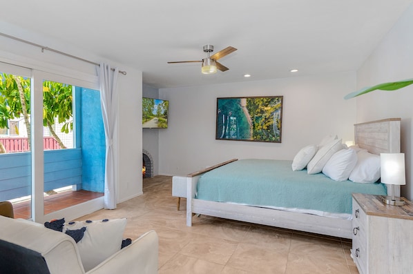 A bedroom with a bed, a ceiling fan, a painting, and a view of the outdoors.