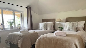 A hotel room with two beds, each with a matching headboard and pillows. There is a window with a view of greenery outside, and a small table with a lamp and a plant. - Kings Cottage - Heart of the Deverills (Warminster)