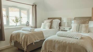 Une chambre avec deux lits, une fenêtre avec des rideaux, une table de chevet avec une lampe et un vase rempli de fleurs.