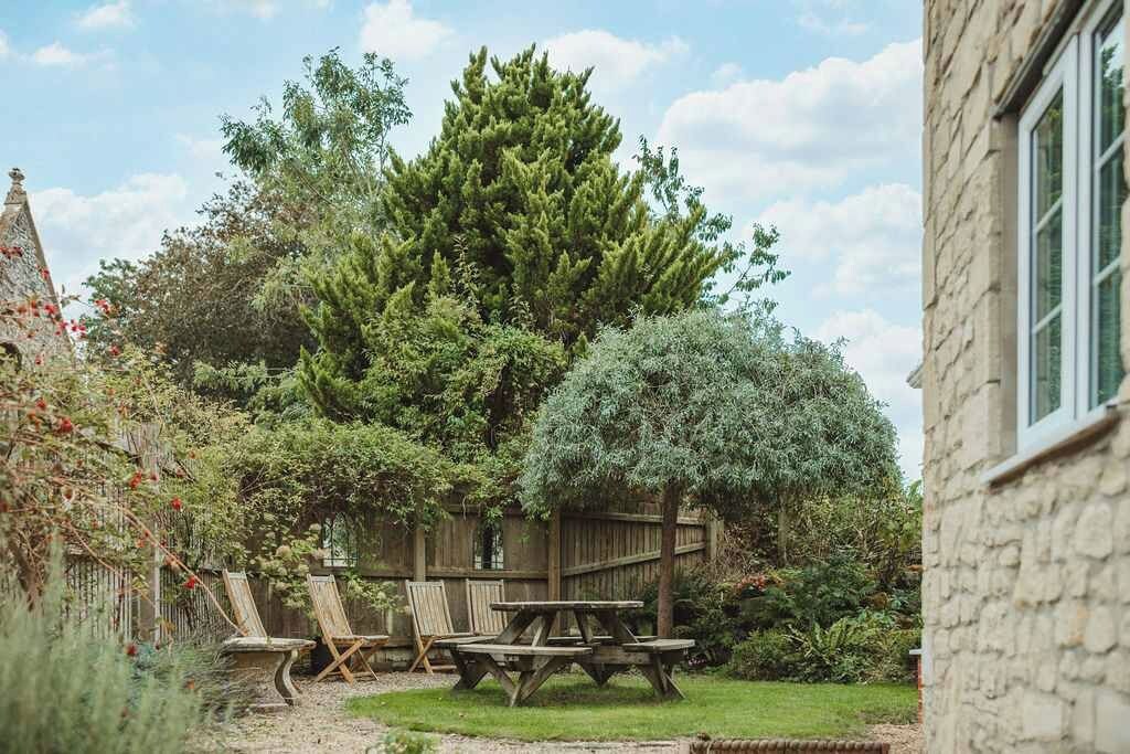 Ein Garten mit einem hölzernen Picknicktisch und Stühlen, umgeben von verschiedenen Bäumen und einem Steinhaus.