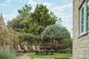 Un jardín con una mesa y sillas de picnic de madera, rodeado de árboles y un edificio de piedra.