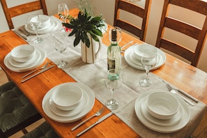 Una mesa de comedor de madera dispuesta con platos blancos, vasos y cubiertos, junto con una botella de vino y un florero con flores.