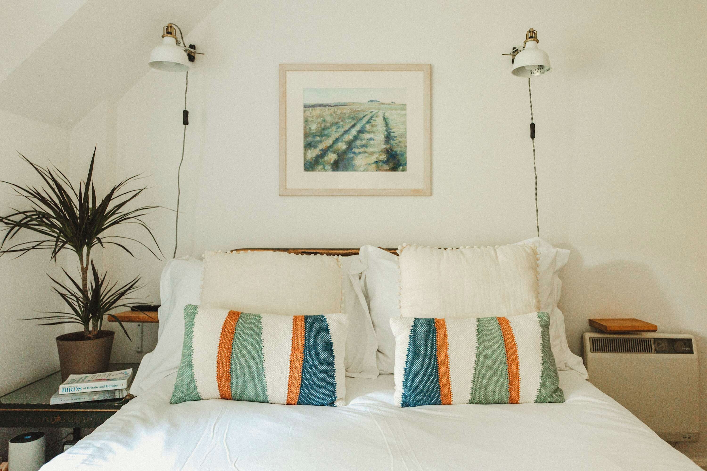 A bedroom with a bed, two bedside lamps, a framed picture on the wall, a potted plant, and a book on a nightstand.