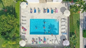 An aerial view of a swimming pool area with sun loungers and umbrellas, surrounded by greenery and a building. - Oceangoing Condo With Pool and Steps to Beach and Food (Marco Island)