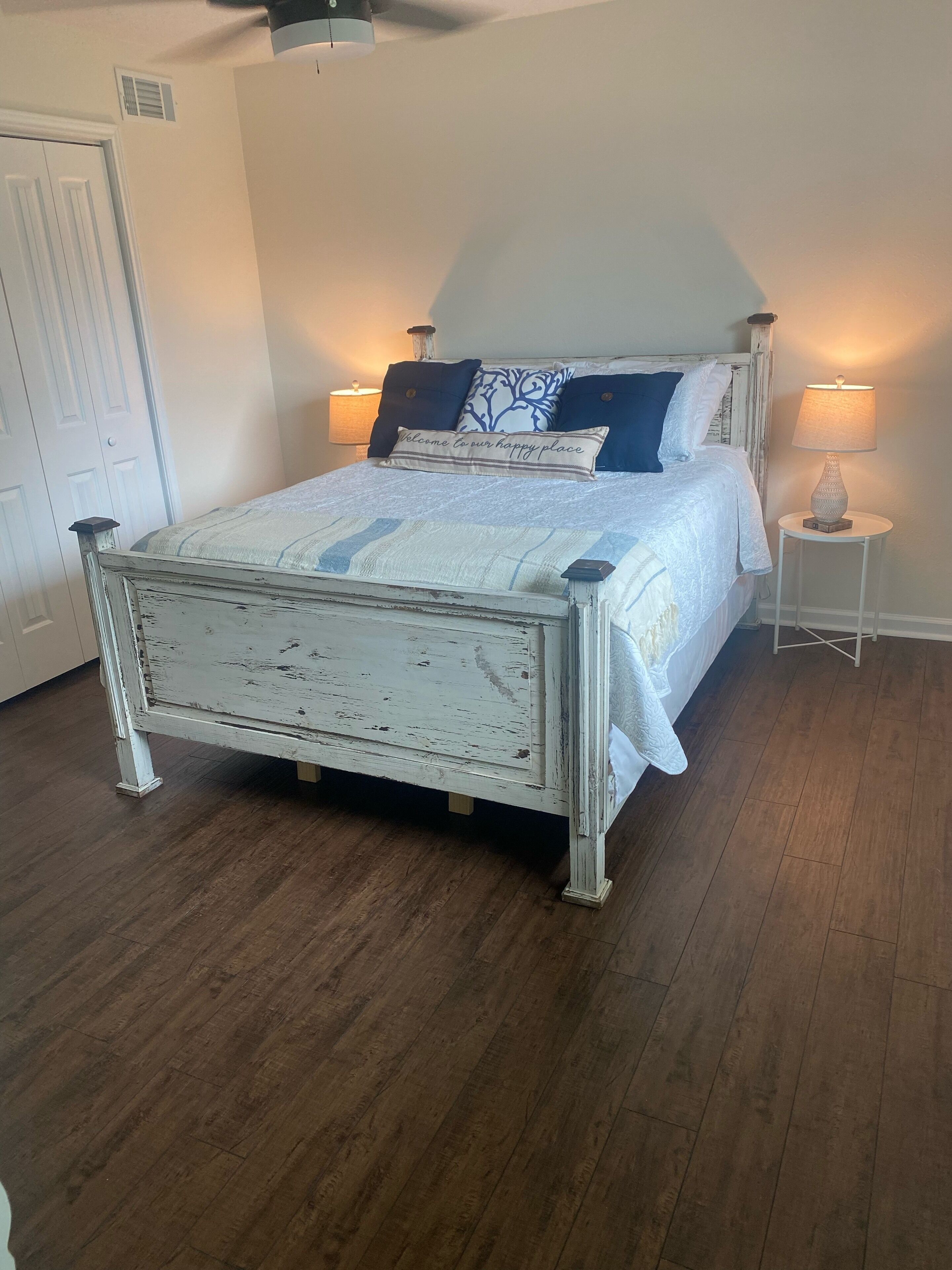 A bedroom with a white, distressed wooden bed, a bedside table with a lamp, and a door.