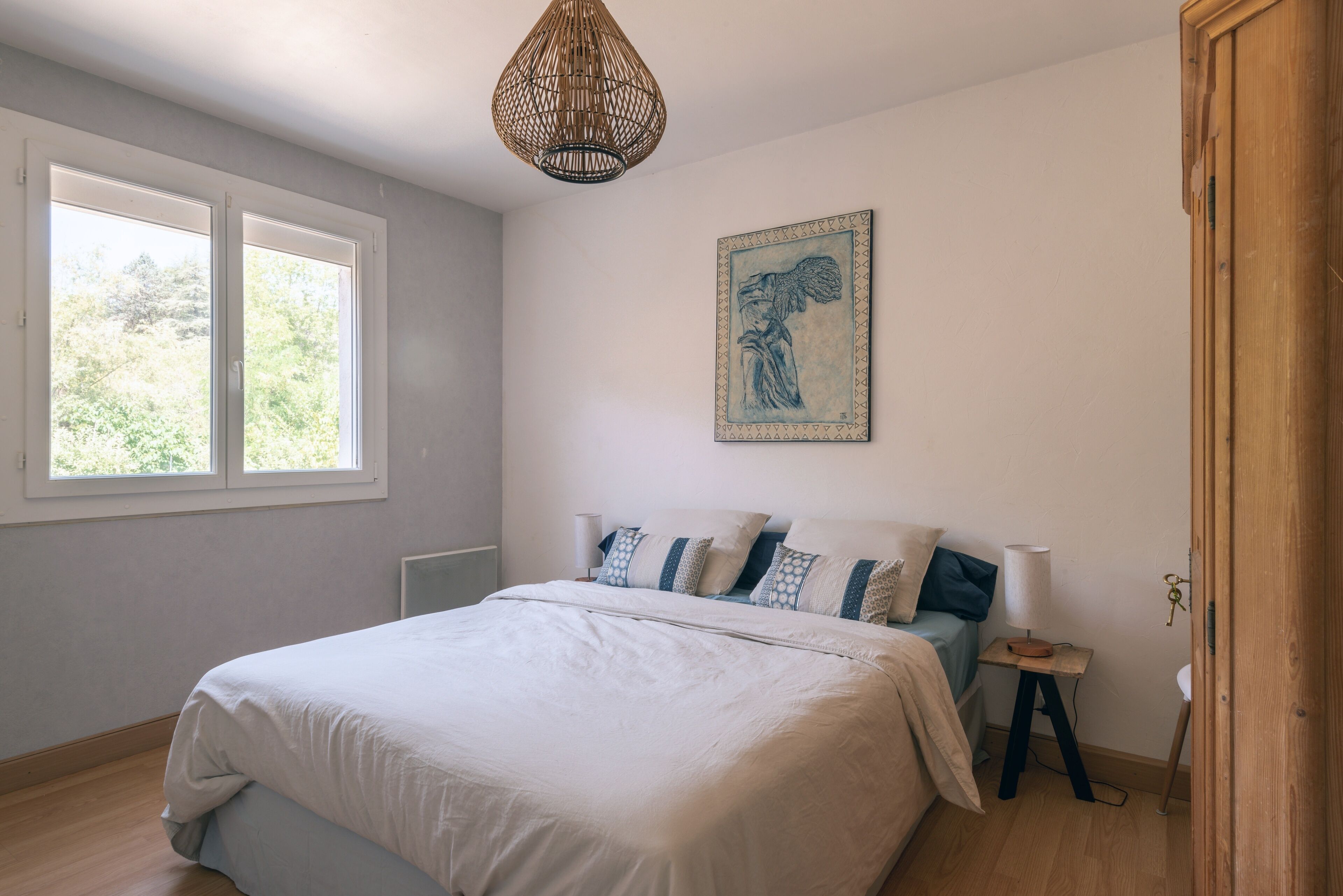 Un dormitorio con una cama grande, una mesita de noche con lámpara, una ventana y una obra de arte en la pared.