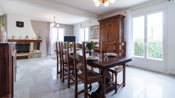 Un comedor con una mesa de madera, sillas y una araña. Se ve un televisor y un armario al fondo.