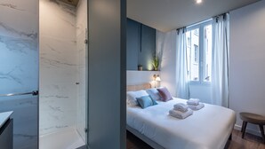 A modern hotel room with a marble shower, a neatly made bed with pillows, a bedside table, and a window with curtains.