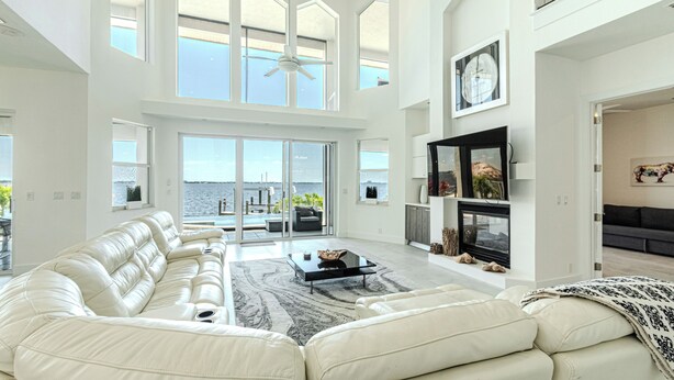 A spacious living room with a large white leather sofa, a coffee table, and a fireplace. There are large windows offering a view of the outdoors.