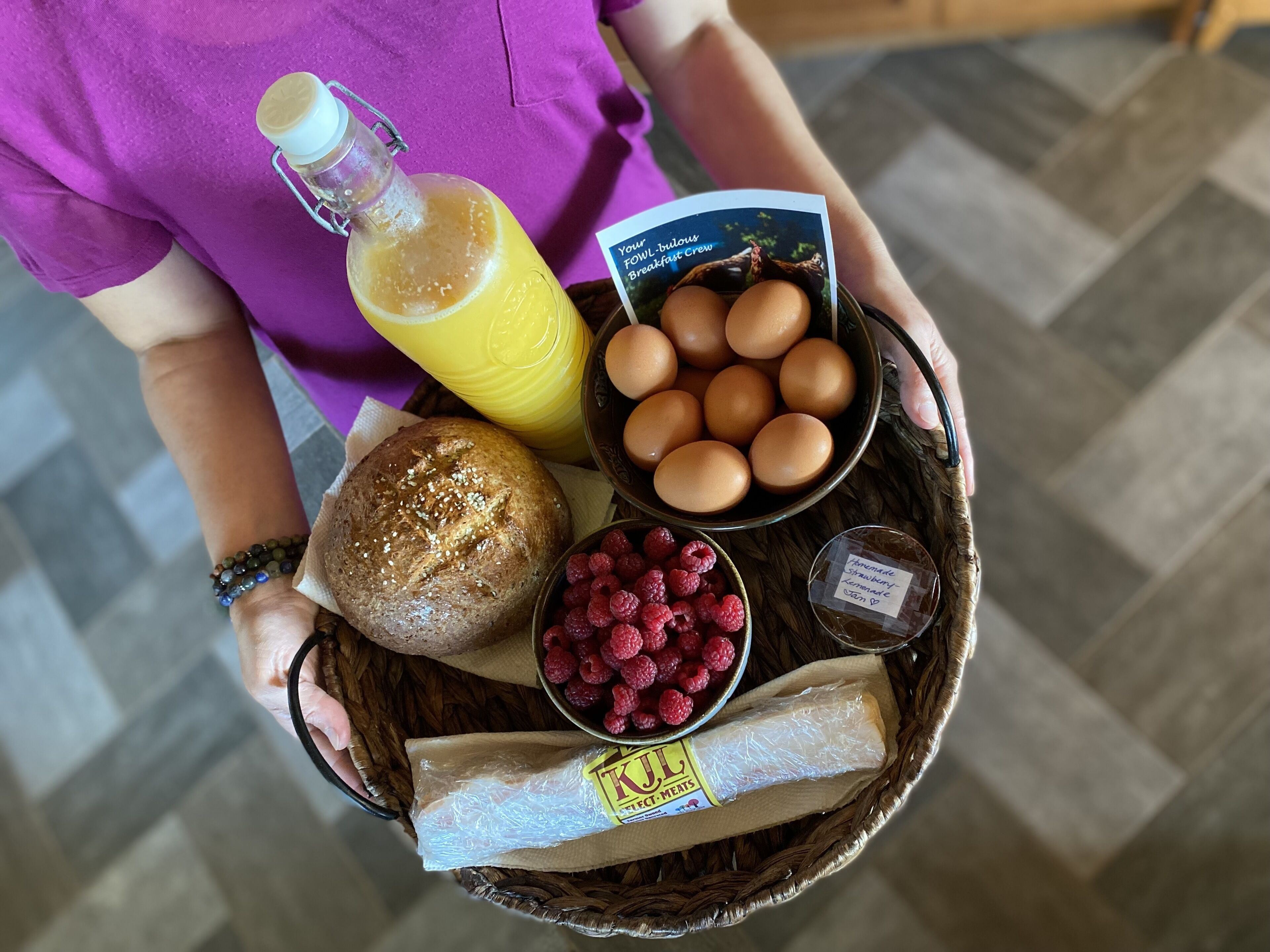 A person holding a basket with bread, eggs, raspberries, a loaf of meat, and a bottle of juice.