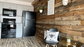 Una cocina con un refrigerador negro, un microondas y una estufa. Se aprecia una pared de madera con un letrero de bienvenida y una planta en maceta. Se ve una silla con un cojín que dice 'Welcome to our Hive'.