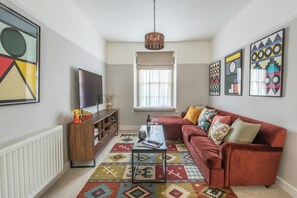 Een woonkamer met een rode bank, een salontafel, een televisie, een kamerkleed met patroon en kunstwerken aan de wanden.