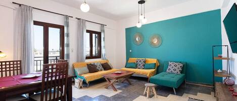 A living room with a sofa set, a dining table, and chairs. There are two windows with curtains, a teal accent wall, and a small stool.