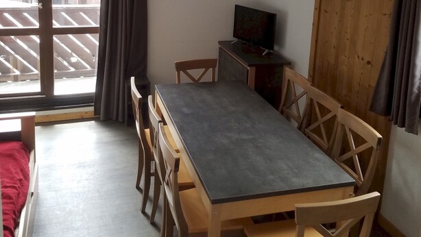 Ein kleiner, gut beleuchteter Raum mit einem Holzespeisetisch und Stühlen, einem Fernseher auf einer Holzkommode und einem großen Fenster mit Vorhängen.