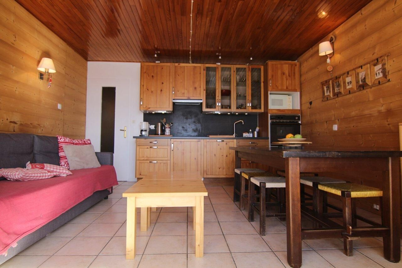 A cozy cabin interior with a wooden ceiling, a sofa, a dining table, and a kitchen area with wooden cabinets and appliances.
