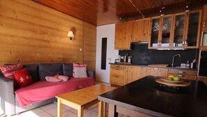 L’intérieur chaleureux d’un chalet, avec un décor en bois, un canapé agrémenté de coussins rouges, une table en bois et un coin cuisine doté de meubles en bois et d’un panier de fruits.