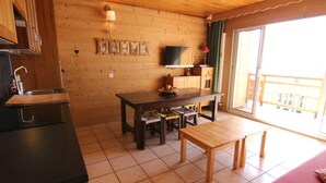 Une chambre confortable, aménagée dans le style d’un chalet, avec un intérieur en bois, un coin repas comprenant une table et des chaises, un téléviseur fixé au mur et une grande fenêtre donnant sur l’extérieur.