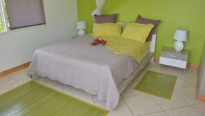 Una cama bien hecha con colcha gris y almohadas amarillas, una mesita de noche con lámpara, y una alfombra verde en el suelo.