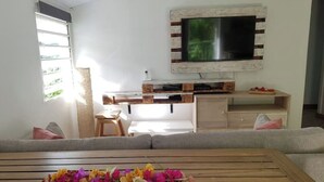 Una sala de estar con un sofá, una mesa de centro de madera con flores, un televisor montado en una pared rústica y un taburete de madera.
