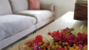 Una bandeja de madera con diversas flores y hojas secas sobre una mesa en una habitación con un sofá blanco y una ventana.