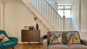 A living room with a staircase, a teal armchair, a grey sofa with colorful pillows, a brown sideboard, and a patterned rug. - Charming Weaver's Cottage in Historic Frome (Frome)