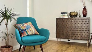 A teal armchair with a colorful cushion, a wooden cabinet with a scalloped design, and a potted plant. - Charming Weaver's Cottage in Historic Frome (Frome)