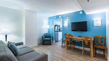 Un salon avec un canapé, une table à manger et des chaises, un téléviseur, et un coin cuisine visible à travers une porte ouverte.